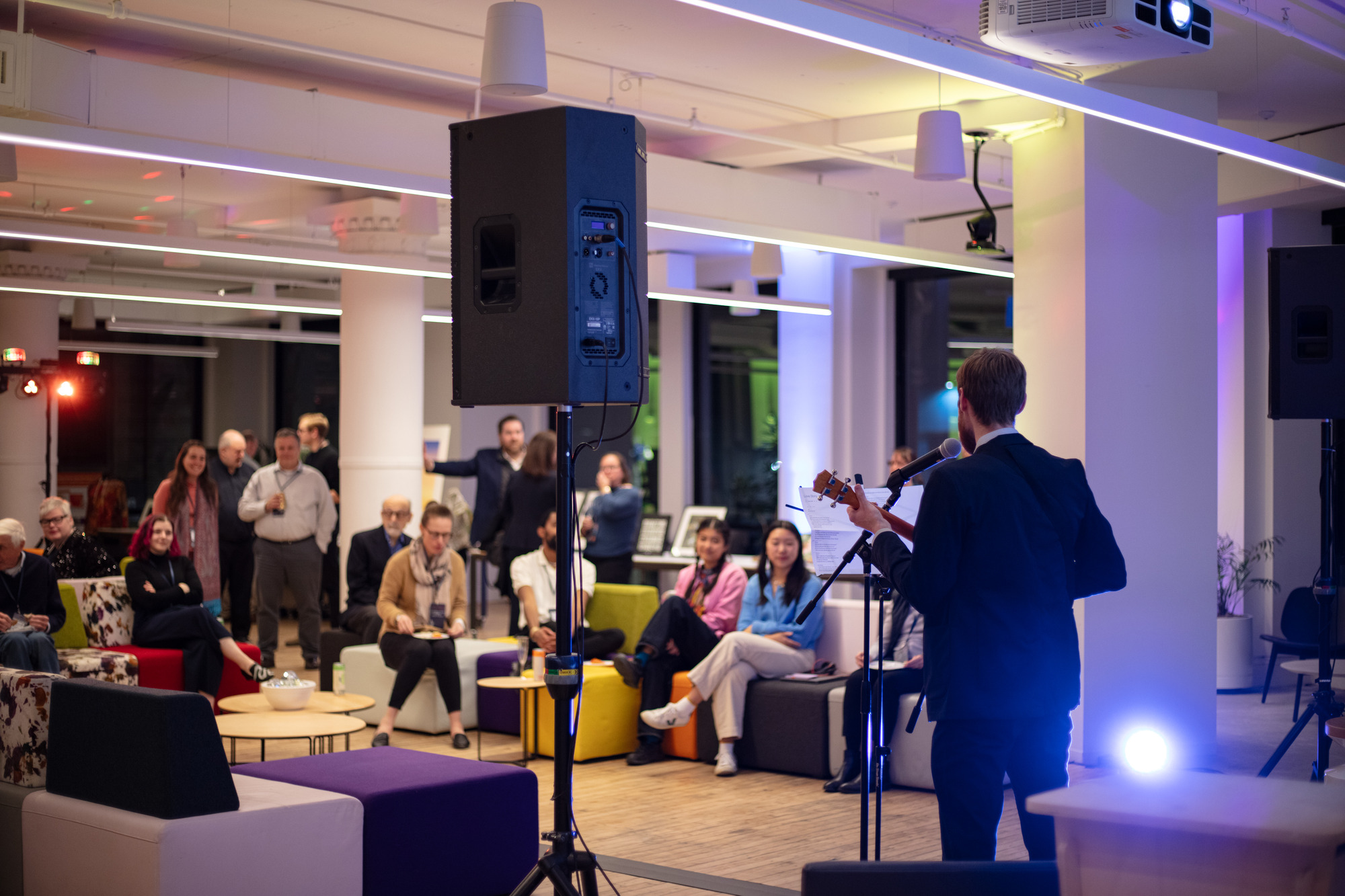 Photo from behind a performer with a mic and ukulele, singing to a crowd in the background