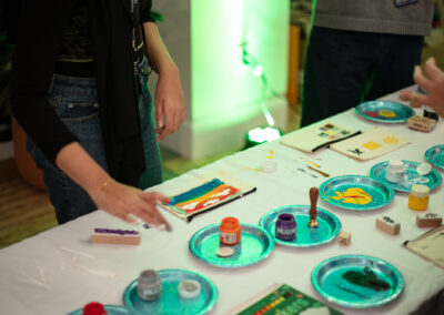 Photo of the Celebration of Design Pencil Case Decorating Station, with paint and stams on plates in the center of a table, and decorated canvas pencil cases drying around the edge.